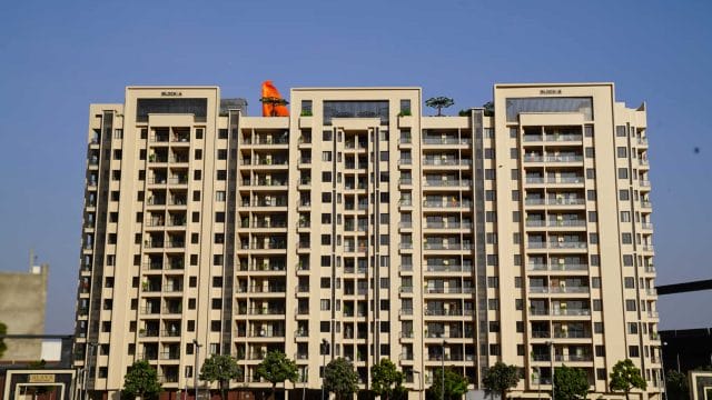 Beige modern residential building with multiple balconies and lush greenery in front.